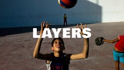 Smiling boy with raised arms looking up at a ball on a sunlit concrete court, with a shadowed wall behind him