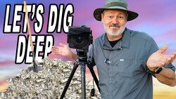 Bearded man in a bucket hat shrugs beside a tripod-mounted camera in front of a pile of cash and a shovel.