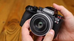 Hands holding a Panasonic Lumix mirrorless camera with a compact silver-rimmed pancake lens facing the viewer