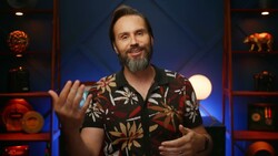 Smiling bearded man in a floral short-sleeve shirt gesturing with both hands in front of a blue-lit shelf set.