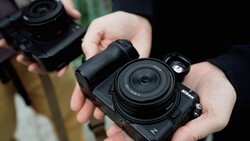 Hands holding a black Nikon Z-series mirrorless camera with a compact pancake lens, another camera blurred in the background