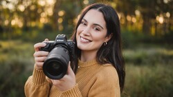 Smiling woman with long dark hair in a mustard sweater holding a Nikon mirrorless camera with a large zoom lens, outdoors in soft light.