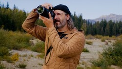 Bearded man in a tan quilted jacket aims a large telephoto lens on a gravel riverbank with conifers and mountains behind.