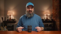 Bearded man in a blue cap and blue shirt pointing at a black camera lens standing on a wooden table
