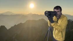 Photographer in a yellow jacket and gloves shooting through a telephoto lens on a tripod above layered mountain ridges at sunrise