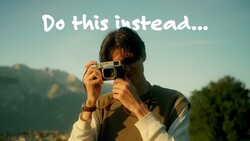 Person in a knit vest raising a silver compact film camera to their eye, with sunlit mountains and trees behind them.