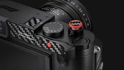 Close-up product shot of a black Leica camera's top plate and carbon-fiber grip, with a red-ringed shutter release and speed dial.