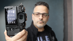 Man in glasses and a black jacket holding up a black mirrorless camera toward the viewer, its rear screen showing menu icons.