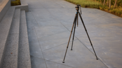 Dark tripod with a fluid video head and panning handle standing on a concrete plaza beside granite steps