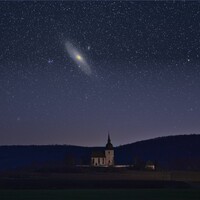 Illuminated church with tall spire beneath starry night sky and Andromeda galaxy
