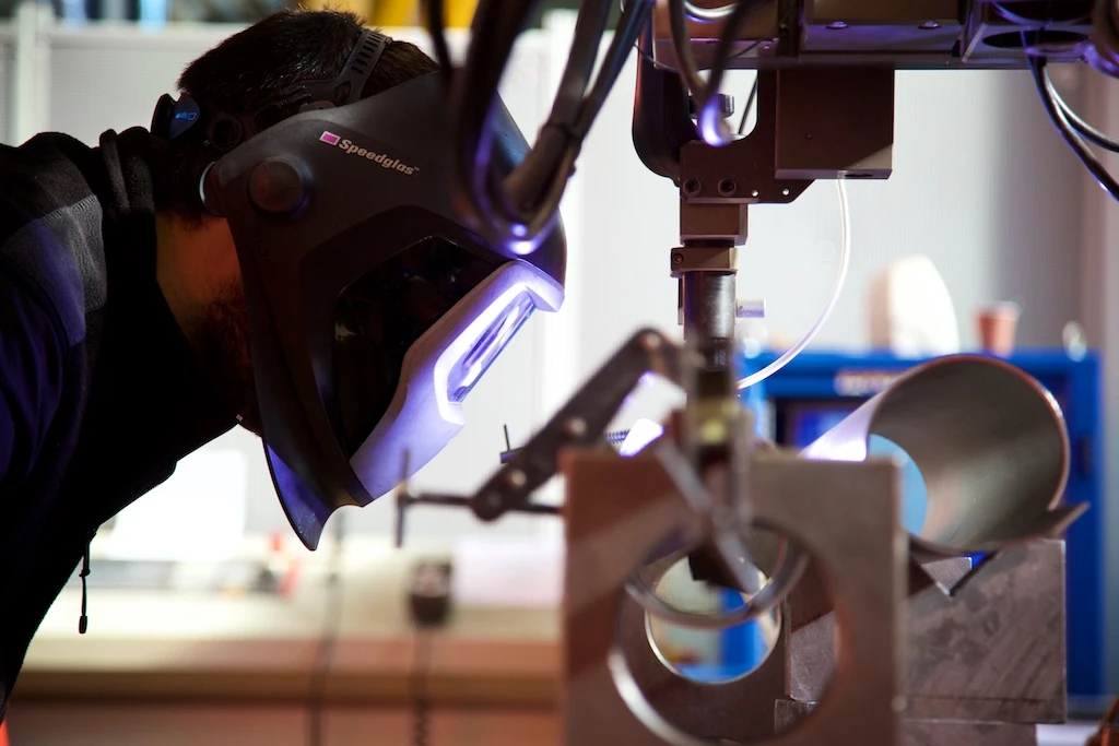 A welder checks the work done by a welding robot at CERN - Photo by Ruben Lammerink