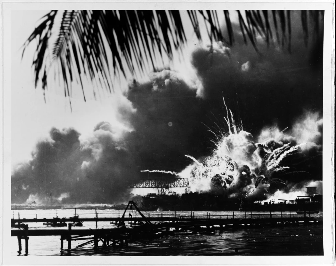 The forward magazine of USS Shaw (DD-373) explodes during the second Japanese attack wave. At right is the bow of USS Nevada (BB-36), with a tug alongside fighting fires. Photographed from Ford Island, with a dredging line in the foreground. U.S. Naval Hi