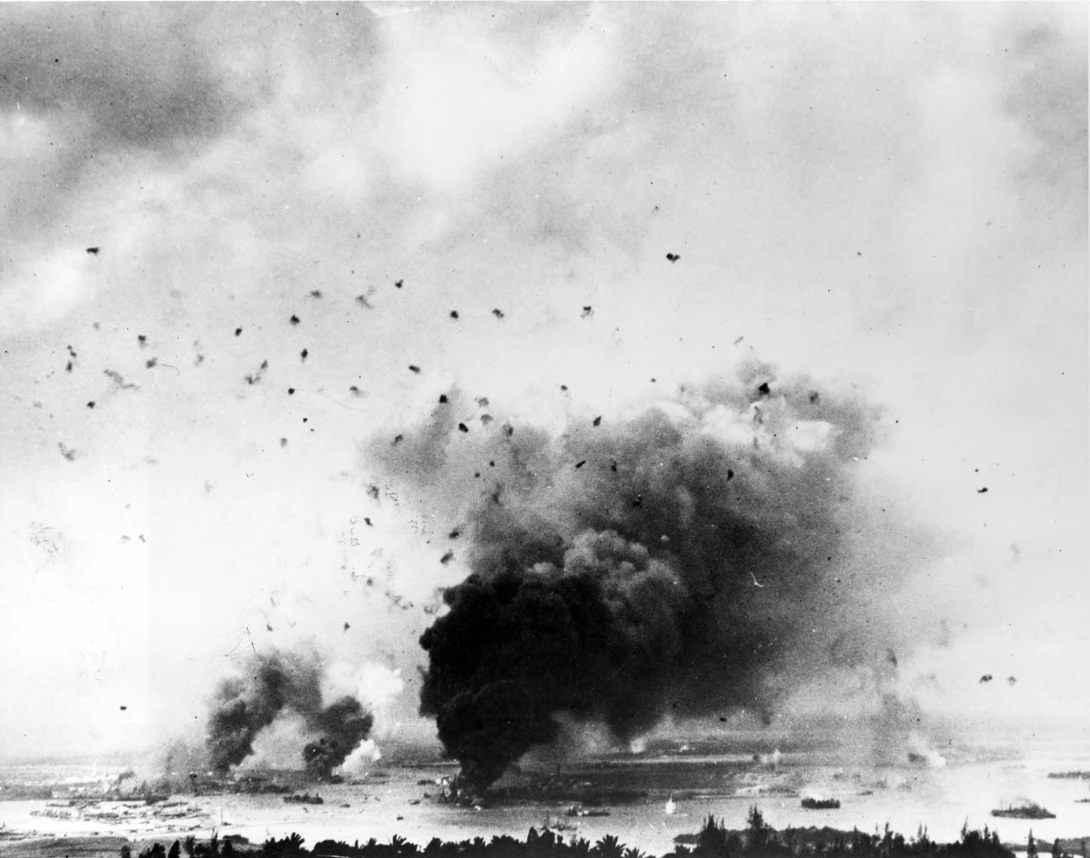 A view of Pearl Harbor looking southwest from the hills towards the north. Taken during the Japanese raid, with anti-aircraft shell bursts overhead. Large column of smoke in lower center is from USS Arizona.