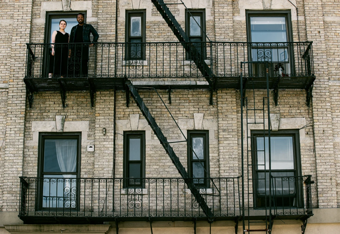A couple stand on their Brooklyn fire escape for a portrait for the Come To My Window project