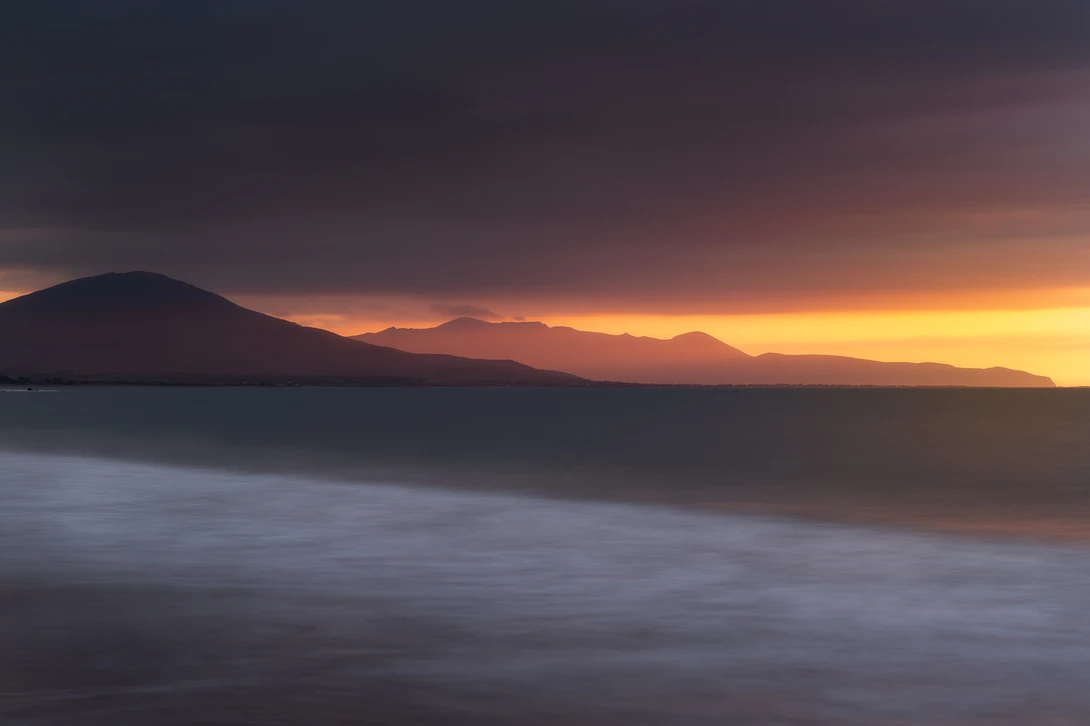 long exposure landscape photo taken on a beach at sunset with mountains in the distance and dramatic light under dark, moody skies 