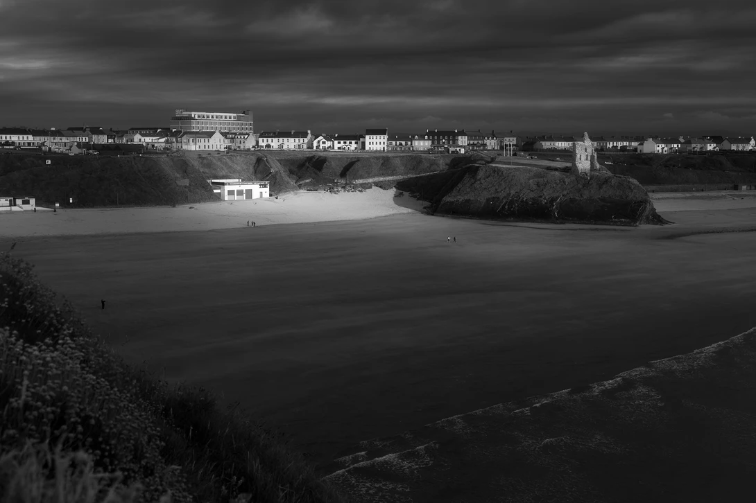 black and white photo of a beach during a dark day shot from cliffs above the water. A break in the clouds illuminates a building on the beach. A number of buildings are in the background.