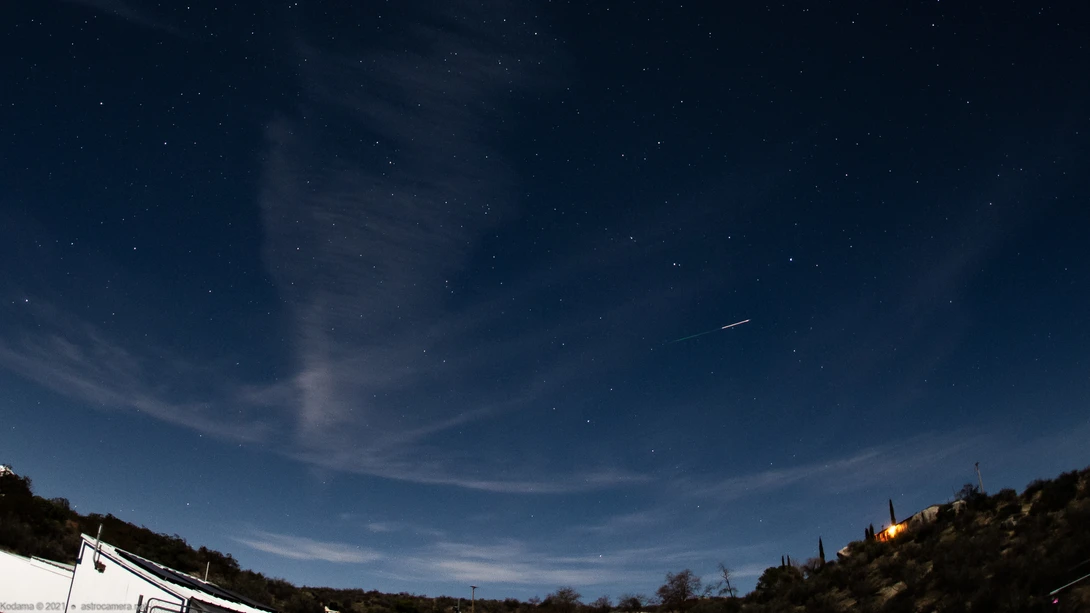 Night sky with stars, thin clouds, and a visible meteor or aircraft streak over a dark landscape with forested hills.