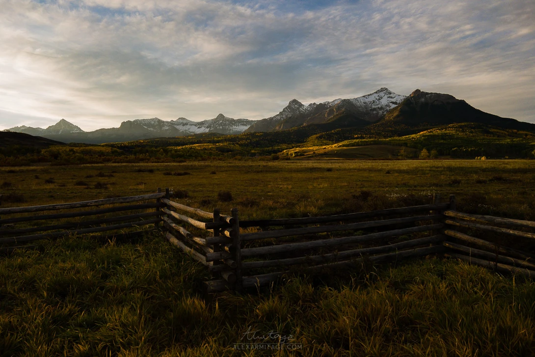 Weathered split-rail fence in golden meadow with snow-capped mountain range in background.