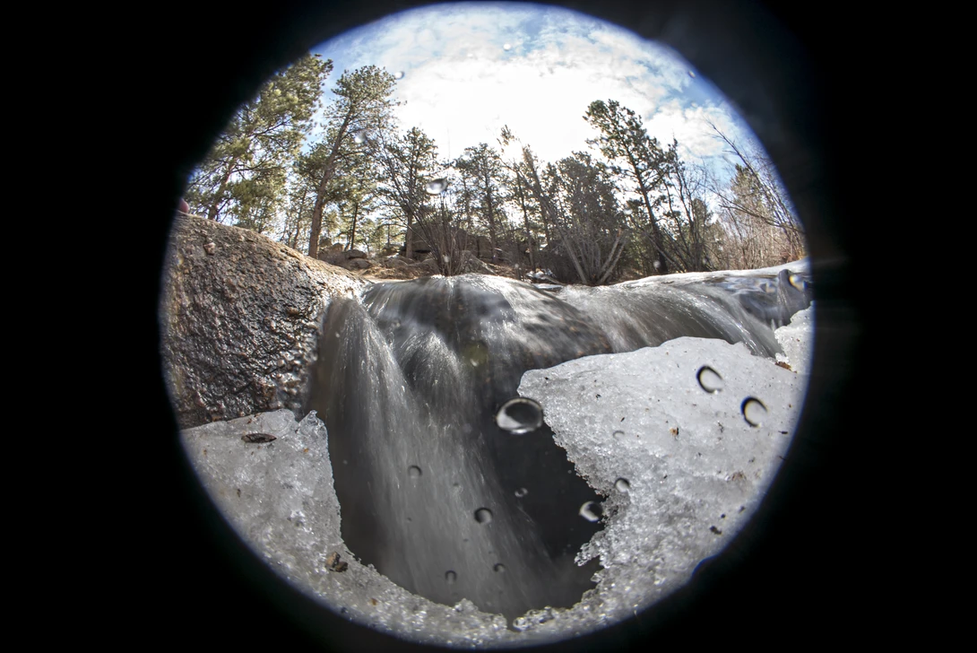 Circular fisheye view of a frozen waterfall with rocky terrain and coniferous trees against a blue sky.