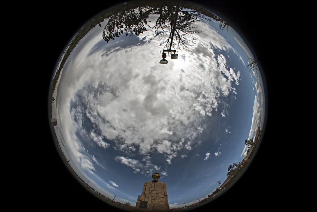 Fisheye photograph of a dramatic sky with towering cumulus clouds and blue sky, framed by tree branches above and a stone structure below.
