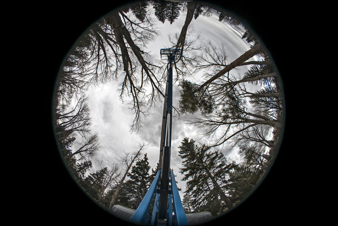 Fisheye view looking straight up at a tall blue pole amid surrounding bare and evergreen trees against cloudy sky.