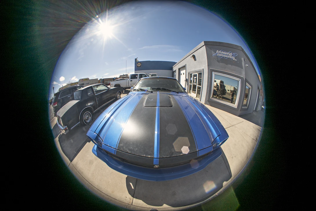 Fisheye view of a blue and black sports car with racing stripes on a dealership lot.