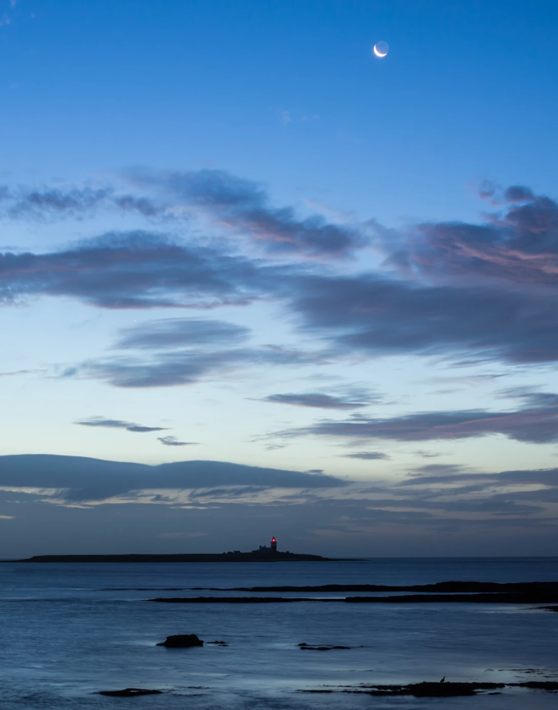 Twilight seascape with crescent moon, layered clouds, and silhouetted rocks in shallow water.