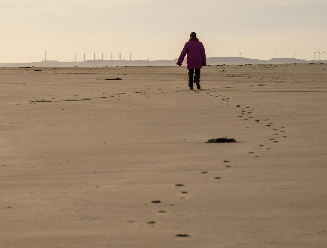 Person in purple jacket walking across flat sandy beach with footprints trailing behind, industrial structures visible on horizon.