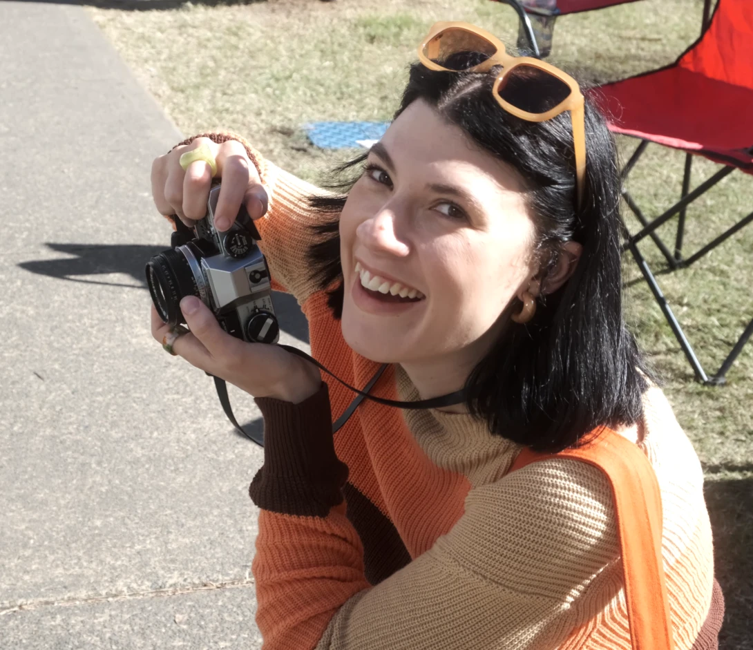 Woman holding a vintage film camera at an outdoor gathering, smiling at the camera.