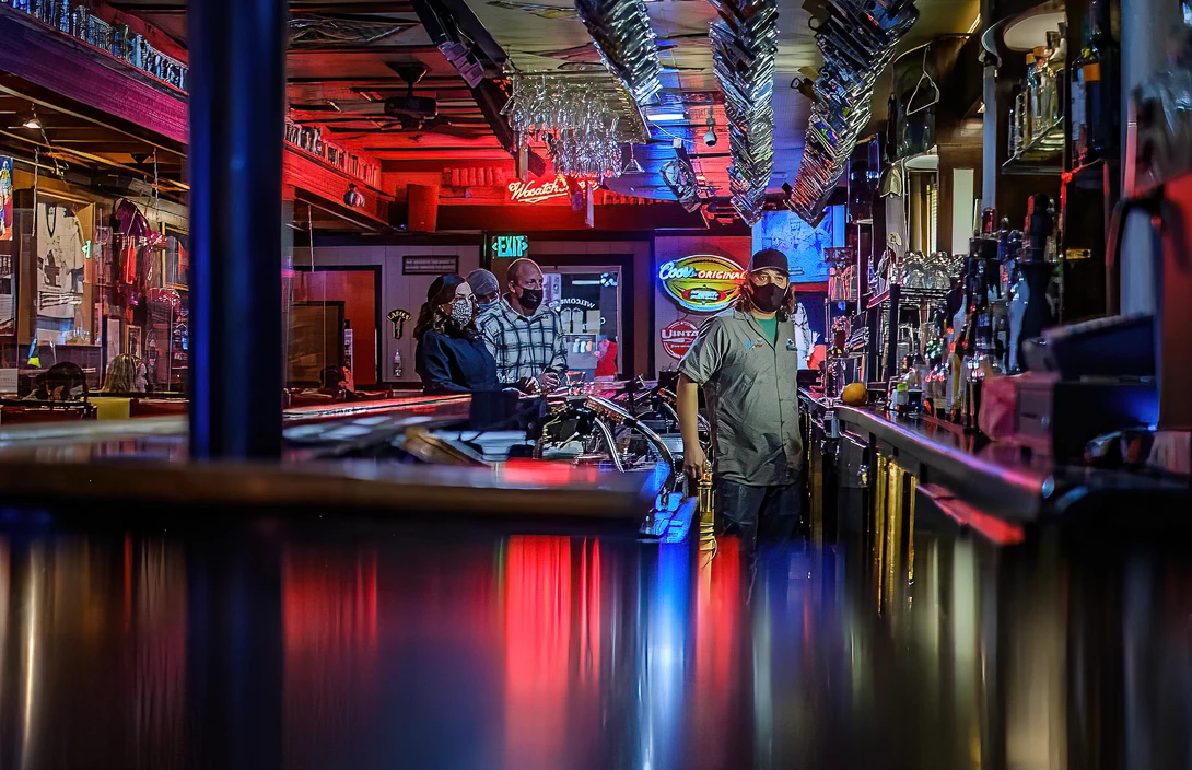 Neon-lit bar interior with red and blue lighting, patrons at counter, motorcycle visible in background.