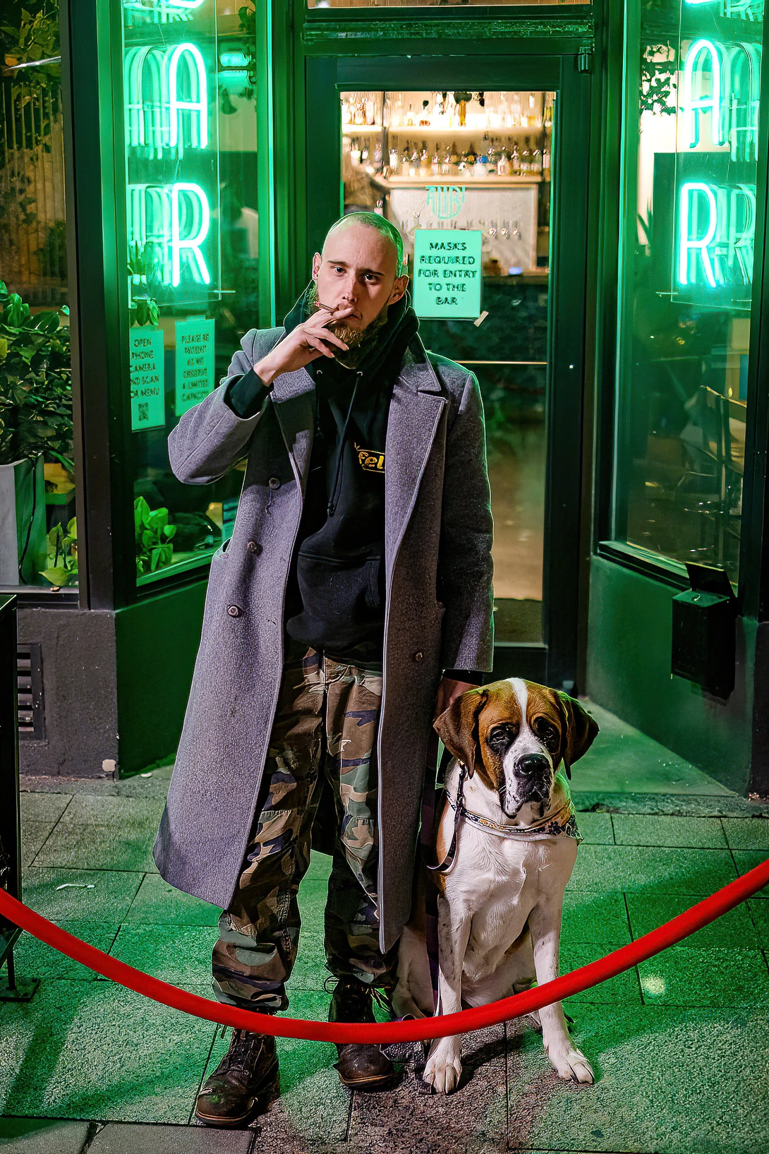 Man in gray coat standing with boxer dog behind red velvet rope in front of neon-lit green storefront.