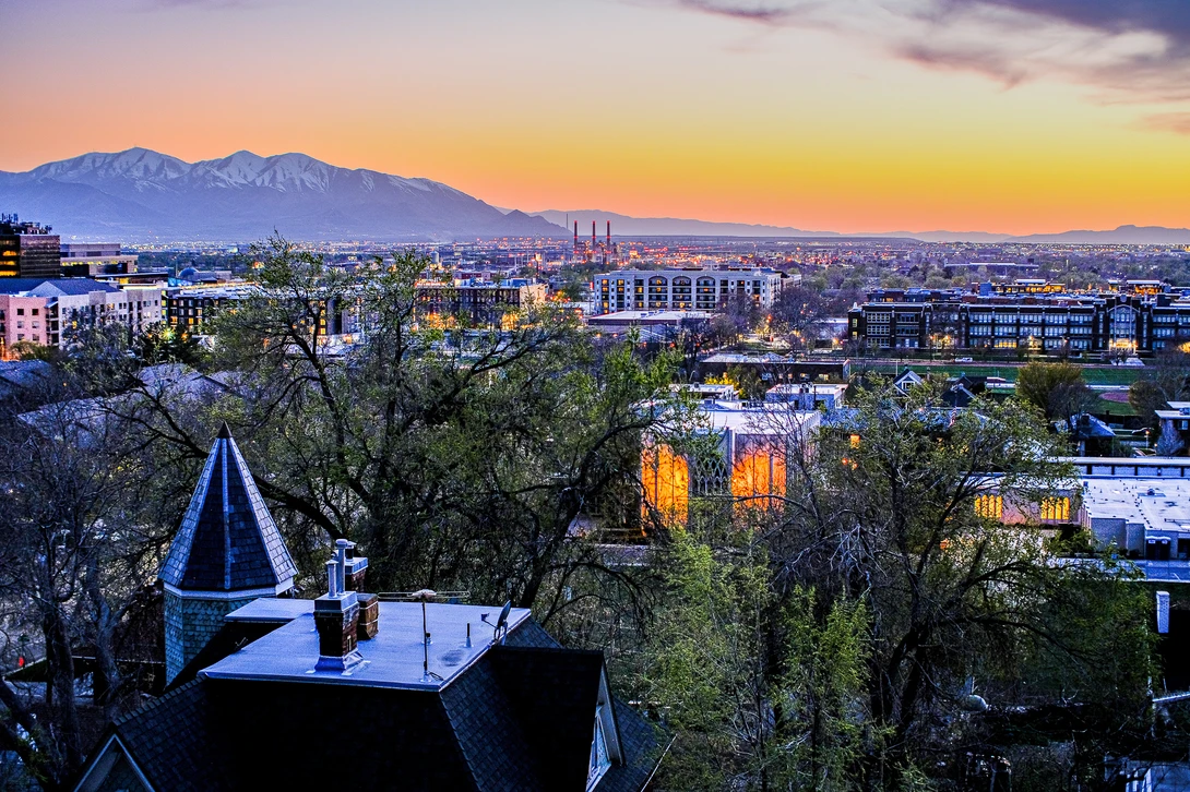 Urban cityscape at twilight with snow-capped mountains in the distance and illuminated buildings below.