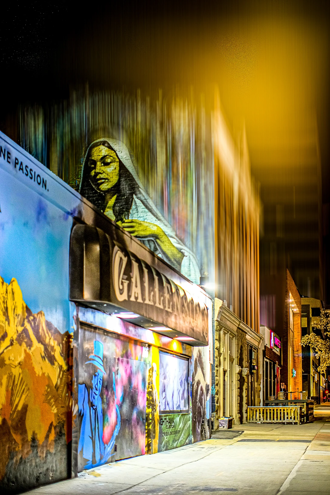 Nighttime street scene featuring large-scale mural art with a woman's portrait in yellow and green tones on a building facade.