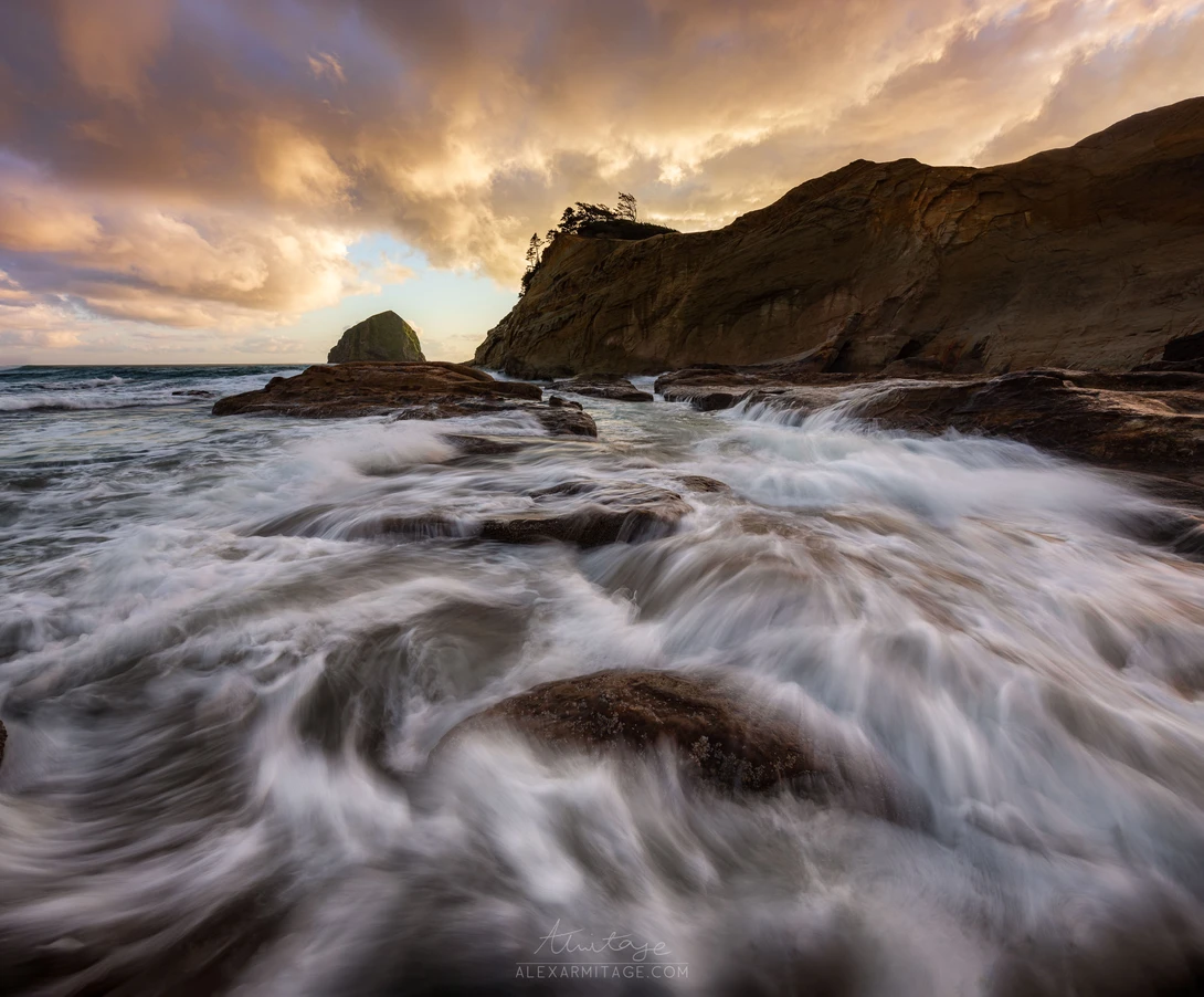 Waves surge across a rocky beach at sunset, with dark cliffs and a sea stack under golden clouds.