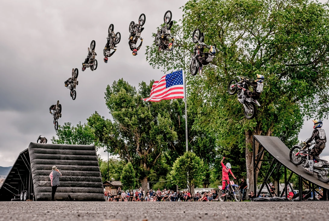 Motocross riders perform aerial tricks on dirt bikes suspended above a ramp during a stunt show.