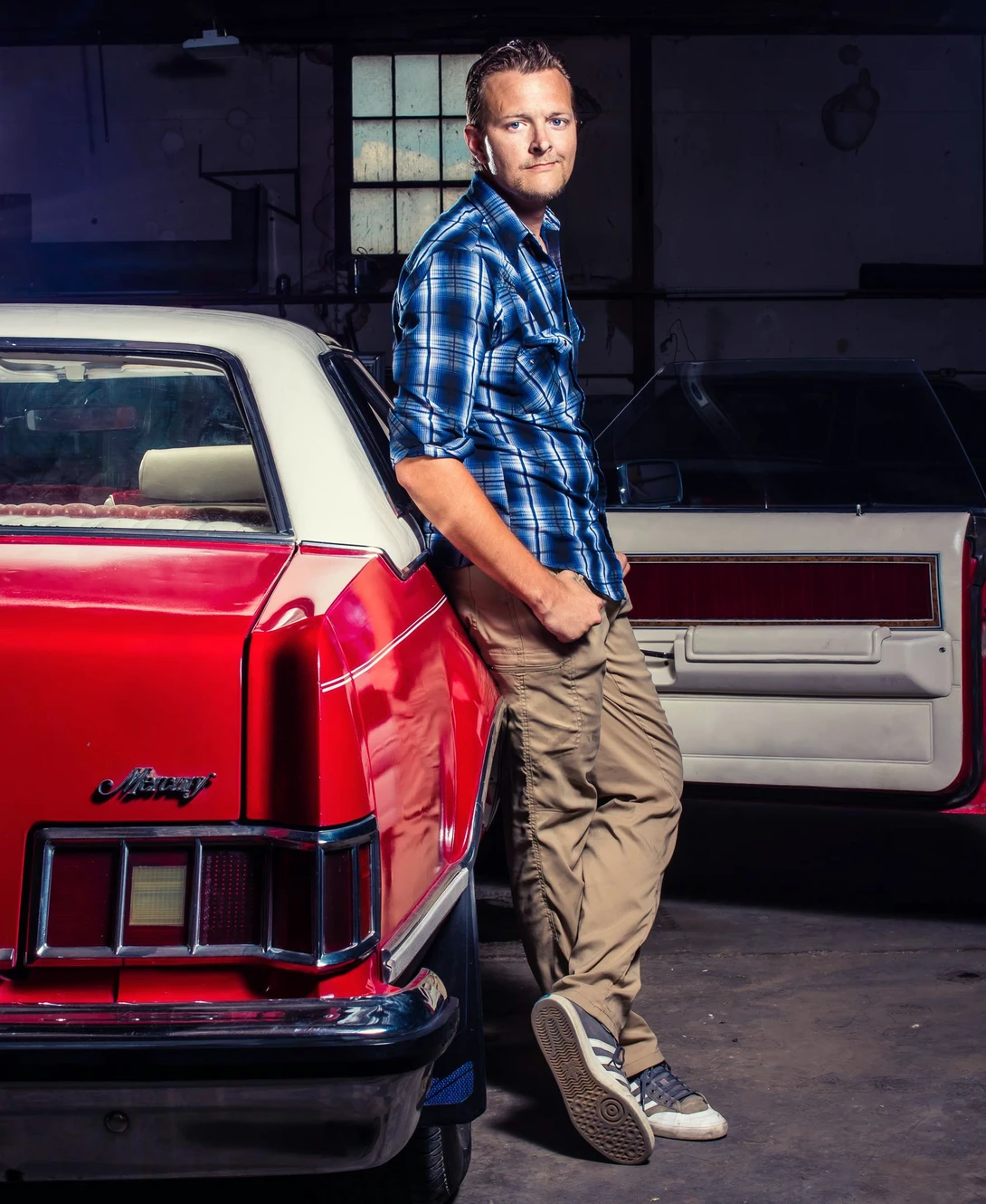 Man in blue plaid shirt leaning against red vintage car in garage setting.