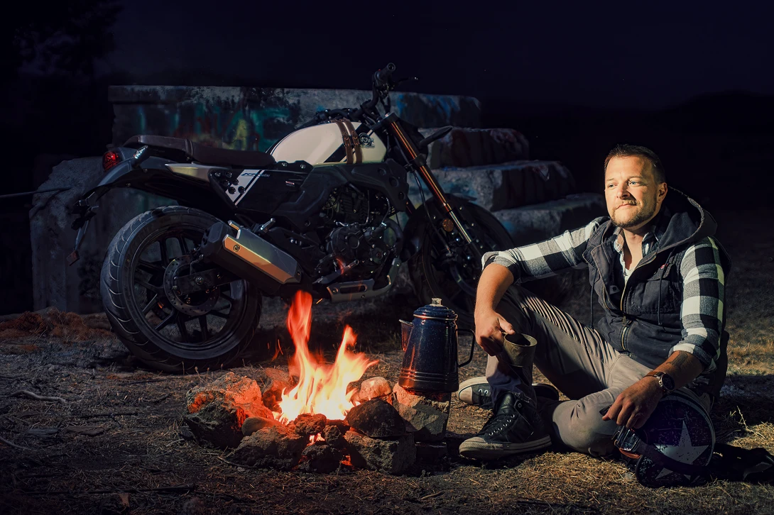 Man crouched by campfire at night with motorcycle and vintage vehicles in background.