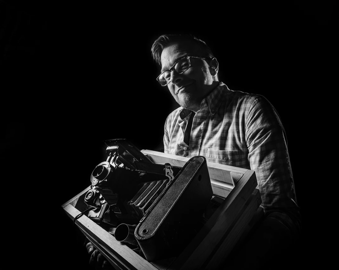 Man with glasses reading from a typewriter in dramatic side lighting against a dark background.