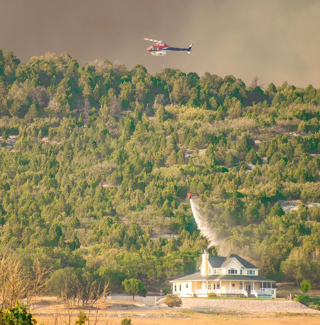 Helicopter dropping water on a forested hillside near a white house during wildfire suppression.