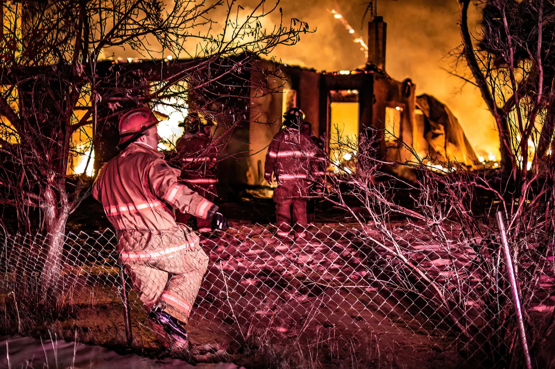 Firefighter silhouetted against intense orange flames while operating from behind chain-link fence.