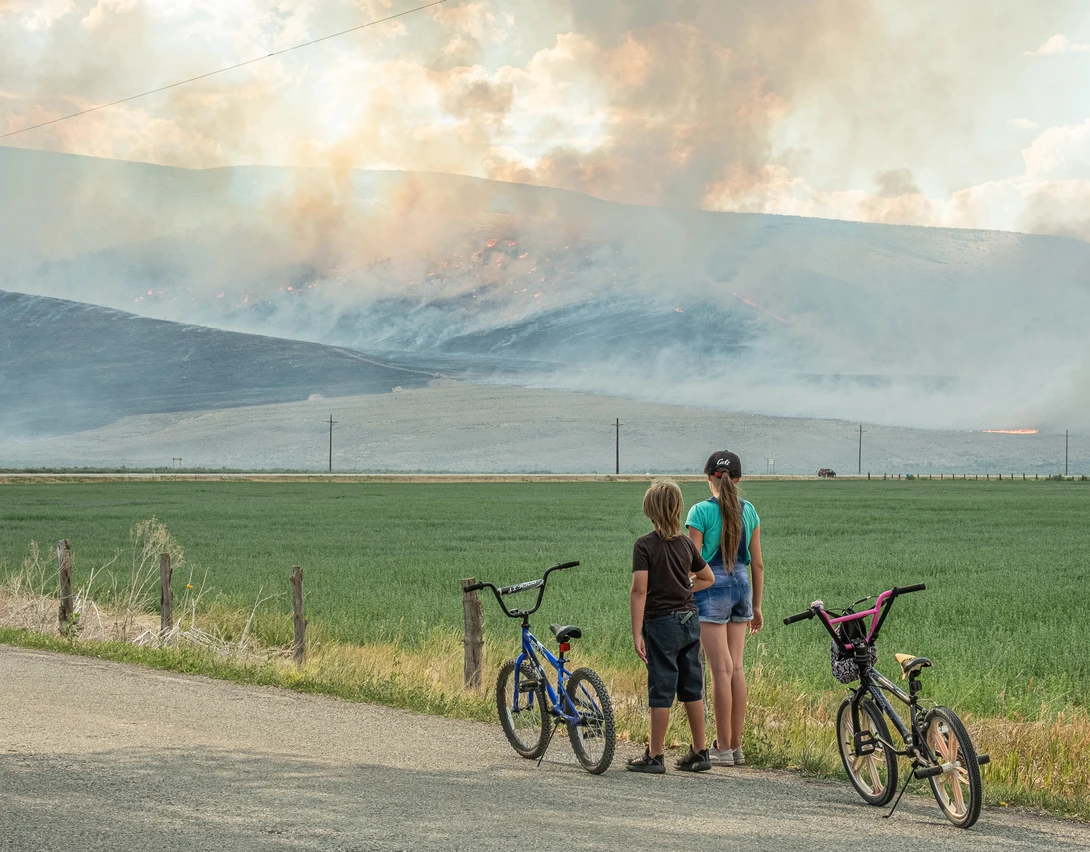 Two cyclists stand with their bikes on a rural road, facing a distant mountain with smoke rising from its peak.