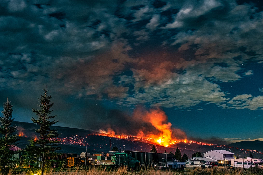 Massive wildfire spreads across mountainside at dusk with intense orange flames and dark smoke against deep blue sky.