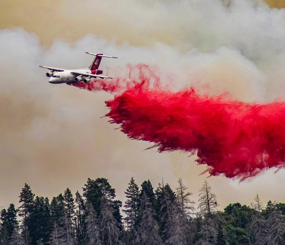 Aerial firefighting aircraft releasing red fire retardant over forested area with smoke and clouds.