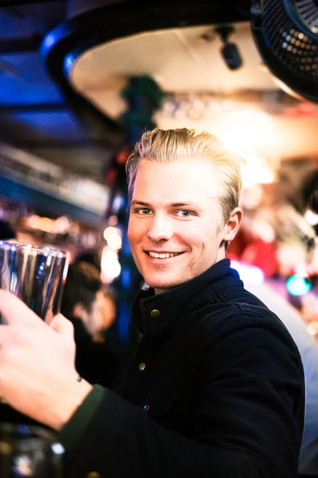 Smiling person with blonde hair holding a glass at an indoor event with blue lighting.