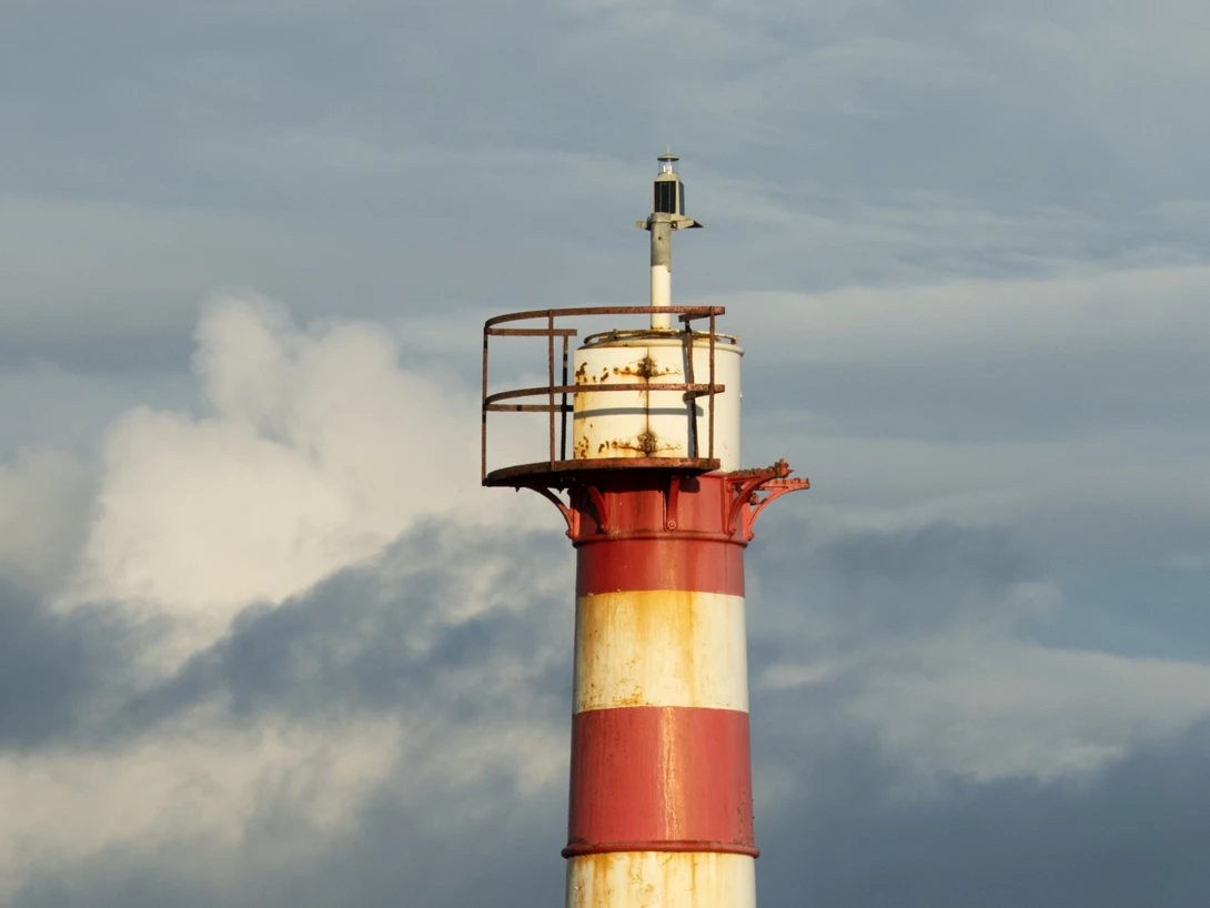 Red and white striped lighthouse tower with golden lantern room against cloudy sky.