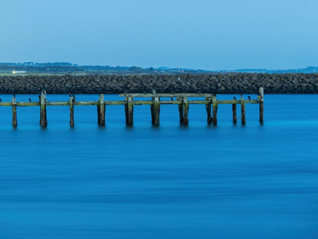 Weathered wooden pilings extending into calm turquoise water with a rocky breakwater and distant shoreline.