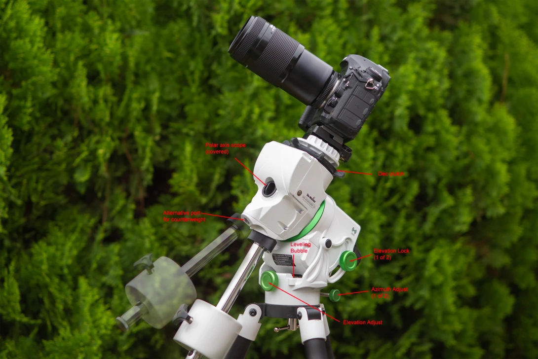 White telescope or spotting scope mounted on tripod with camera attached and labeled components against green foliage background.