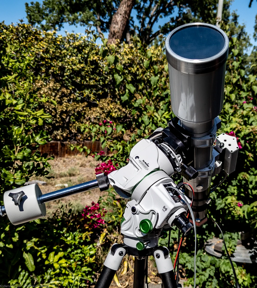 White equatorial telescope mount with long black refractor telescope pointed skyward against blue sky and green foliage.
