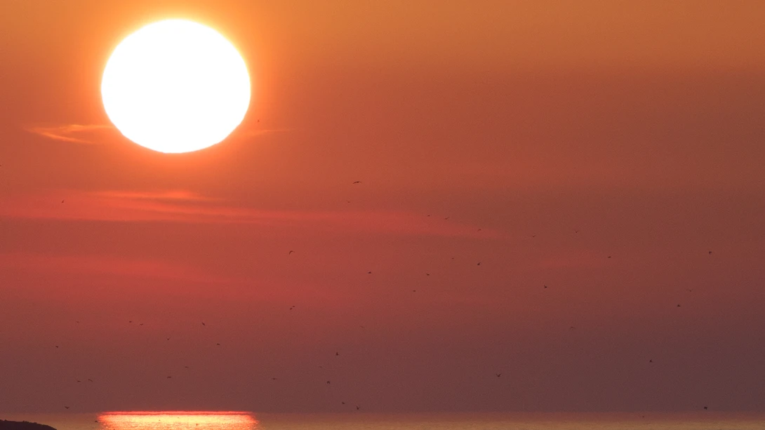 Bright sun positioned in upper left of frame against deep orange sky at sunset or sunrise.