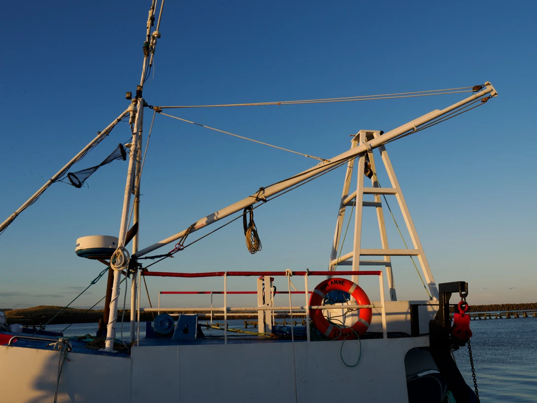 Fishing vessel deck with wooden crane boom and life ring against clear blue sky and calm water.